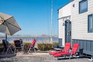 A waterfront outdoor seating area with chairs and tables, a large umbrella, and a building with a door and windows.