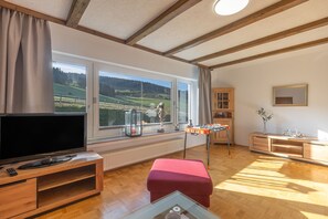 A room with a television, windows with a view of the countryside, a wooden cabinet, and a red ottoman.