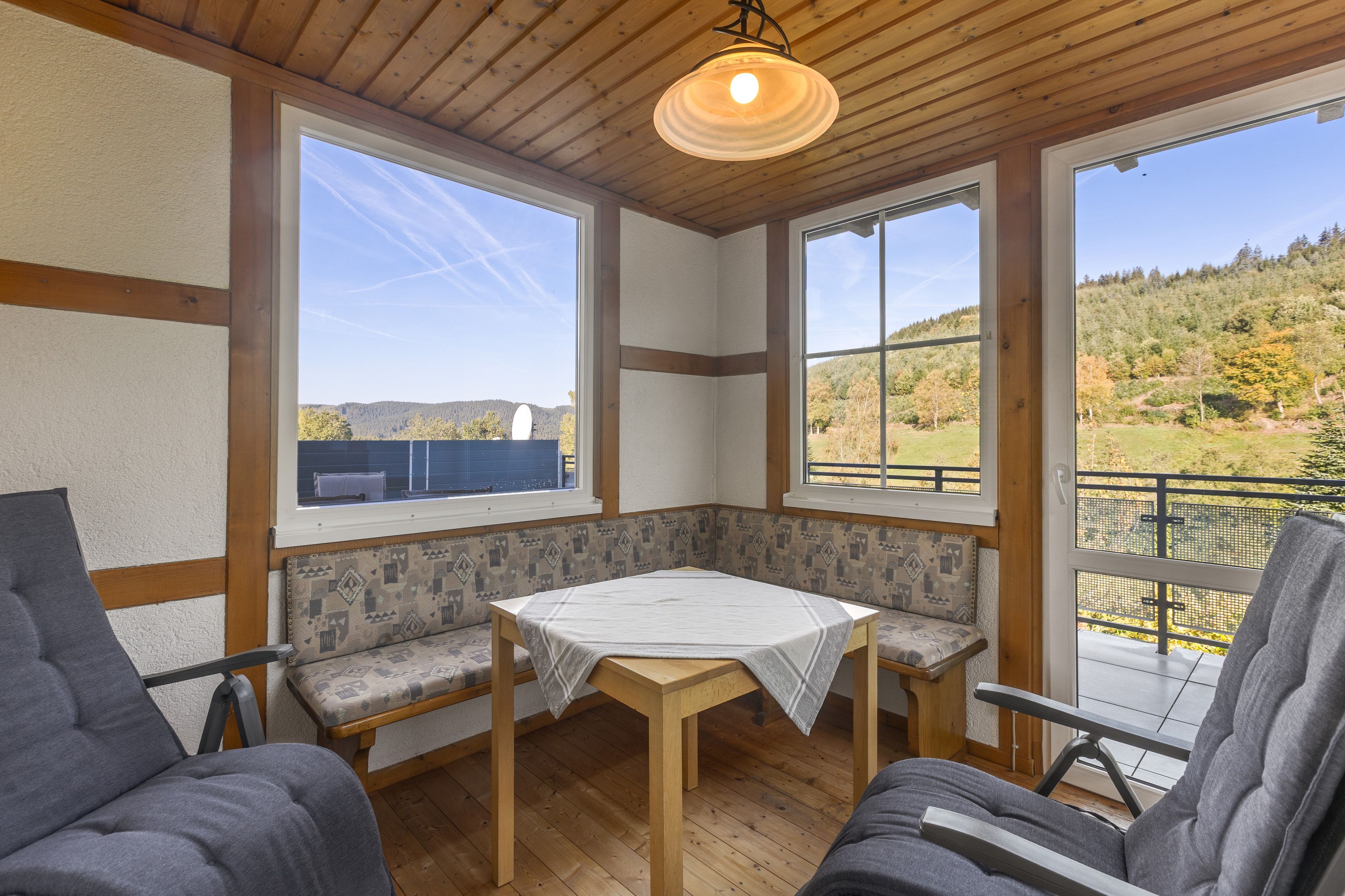 A corner room with a table and two chairs, large windows with a view of a forested landscape, and a wooden ceiling.