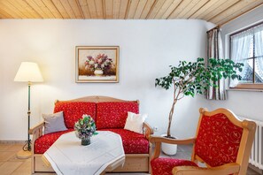 Una stanza accogliente con soffitto in legno, un divano rivestito con tessuto rosso a motivi, un tavolo con tovaglia bianca, una lampada da terra, una pianta in vaso e una finestra con tende.