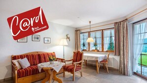 A cozy room with a wooden sofa, a dining table, and a chair. There are framed pictures on the wall, a lamp, and a window with curtains. - Holiday Apartment 'Type B Apartment 2' with Mountain View, Shared Terrace and Wi-Fi (Feldberg (Schwarzwald))