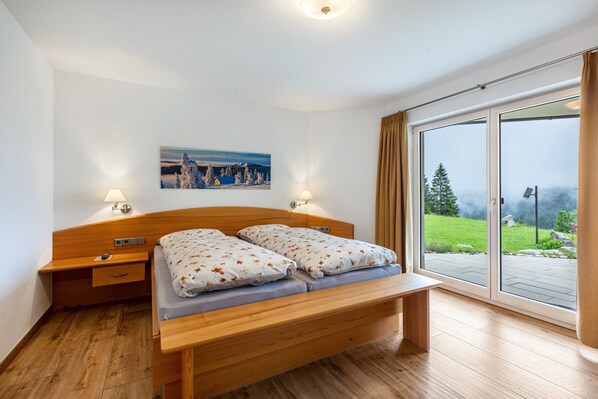 A bedroom with a wooden bed, two bedside tables, a nightstand, a painting on the wall, and a large sliding glass door leading to a garden.