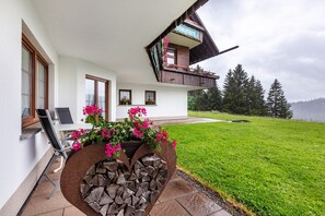 A two-story house with a balcony, a dining area with a table and chairs, and a garden with flowers and a log planter.