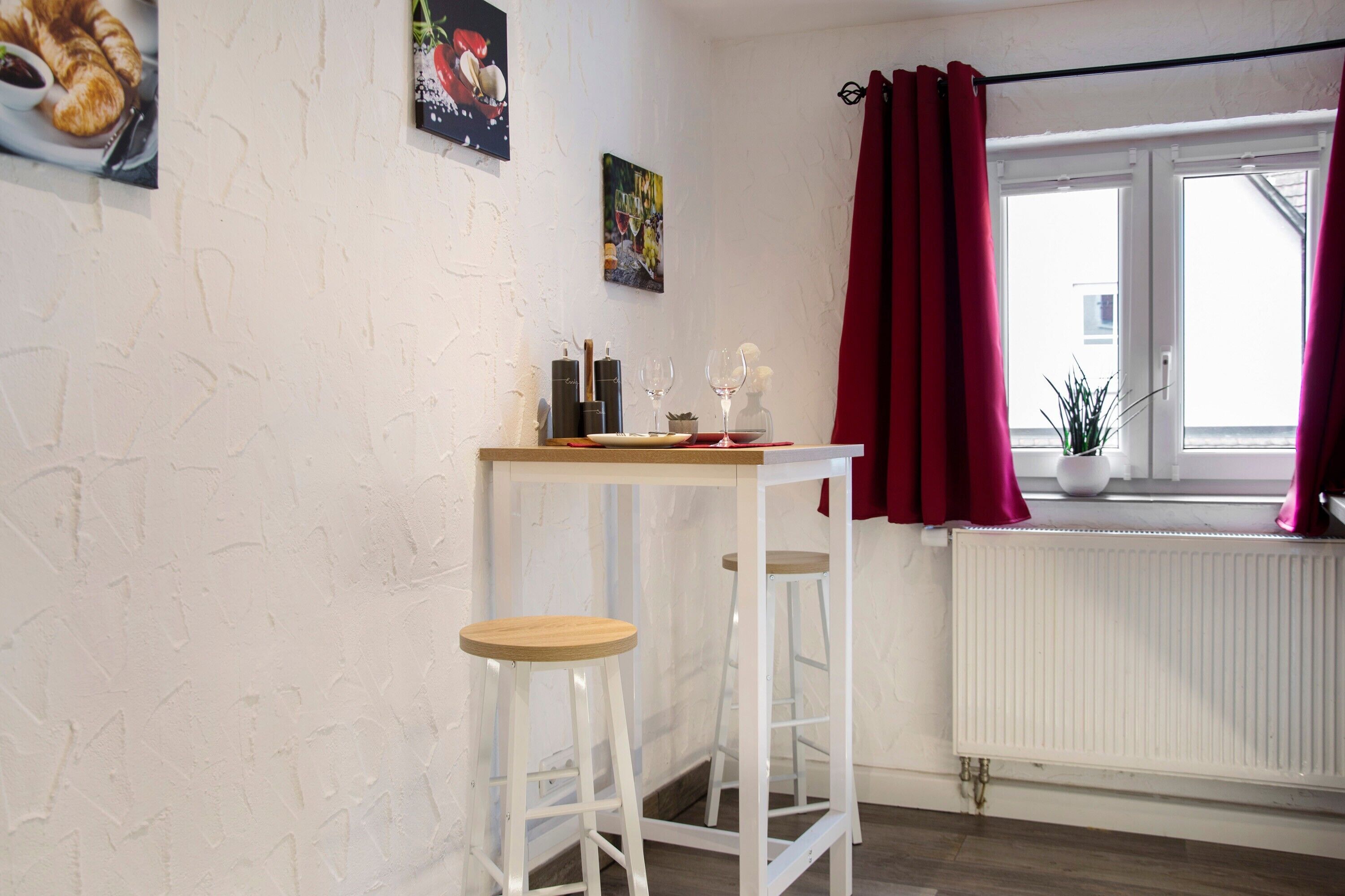 A room with a white textured wall, a window with red curtains, a radiator, and a table with two stools.