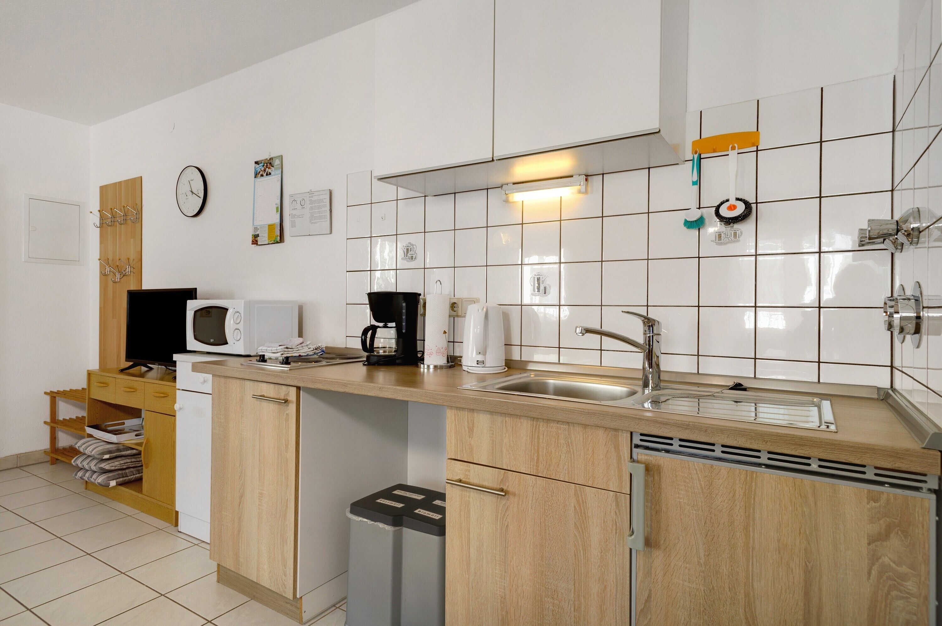 Eine kleine Küche mit Mikrowelle, Kaffeemaschine und Spüle. Ein Holzschrank und ein Mülleimer sind vorhanden.