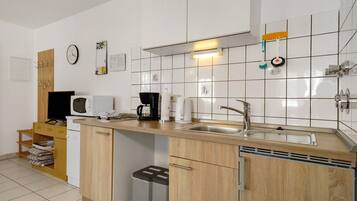 Eine kleine Küche mit Mikrowelle, Kaffeemaschine und Spüle. Ein Holzschrank und ein Mülleimer sind vorhanden.