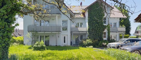 A two-story house with a balcony, surrounded by greenery and parked cars.