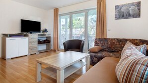 Un salon avec un téléviseur à écran plat, une table basse en bois, un canapé et un fauteuil. Il y a une fenêtre avec des rideaux et un tableau au mur.