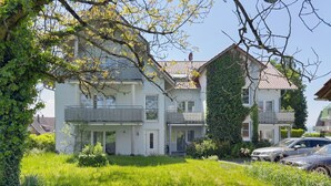 Une maison à deux étages avec un balcon, entourée de verdure et de voitures garées.