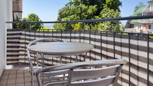 Ein Balkon mit rundem Tisch und Stühlen, einer gestreiften Stoffbarriere und Blick auf einen großen Baum sowie ein Gebäude im Hintergrund.