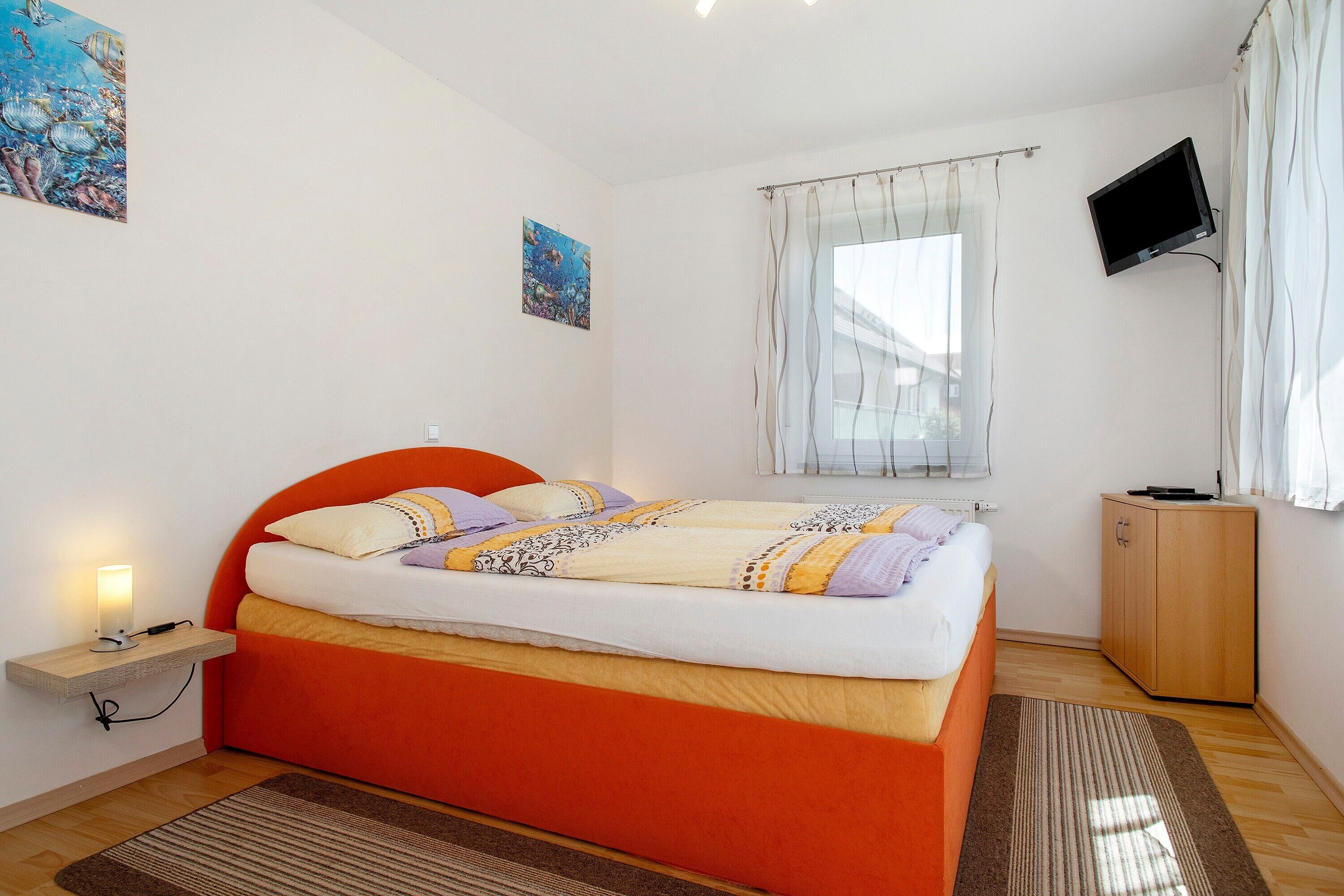 Quarto com cama, mesa de cabeceira com luminária, TV na parede e janela com cortinas.