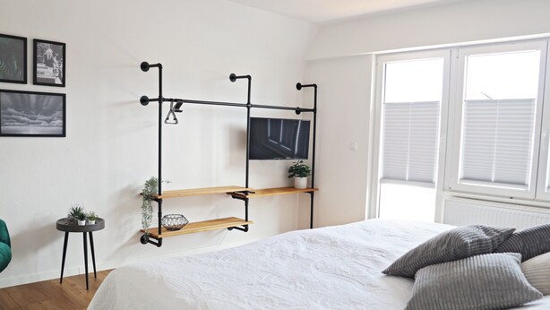 A bedroom with a bed, a TV mounted on a wall, a window with blinds, and a small side table with a plant. - Holiday Home 'Hq16 - Modern' with Private Terrace, Balcony and Wi-Fi (Neuenburg am Rhein)