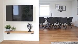 A modern living room with a flat-screen TV, a wooden shelf with plants, and a dining area with a table and chairs. - Holiday Home 'Hq16 - Modern' with Private Terrace, Balcony and Wi-Fi (Neuenburg am Rhein)