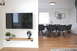 A modern living room with a flat-screen TV, a wooden shelf with plants, and a dining area with a table and chairs. - Holiday Home 'Hq16 - Modern' with Private Terrace, Balcony and Wi-Fi (Neuenburg am Rhein)