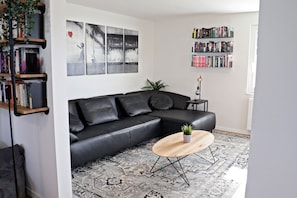 En moderne stue med en sort lædersofa, et rundt træsofabord, et mønstret tæppe, en reol og en vægplante.