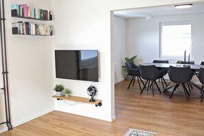 En moderne stue med et fladskærms-tv, bogreoler, et spiseområde med stole og et stort vindue.