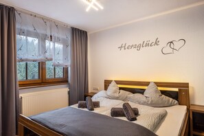 Ein ordentlich bezogenes Bett mit Kissen und Handtüchern, ein Fenster mit Vorhängen sowie eine Wand mit dem Schriftzug 'Herzlich' und einem Herzsymbol.