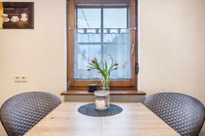 Ein Esstisch mit Blumen in einer Vase, zwei Stühle, ein Fenster mit Vorhang und ein an der Wand hängendes Bild.