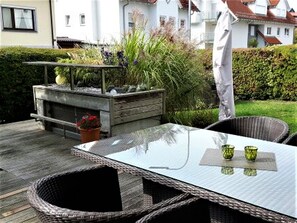 Eine Terrasse im Freien mit Korbstühlen, einem Glastisch und einem kleinen Gartenbereich mit Pflanzen und einem Holzpflanzkasten.