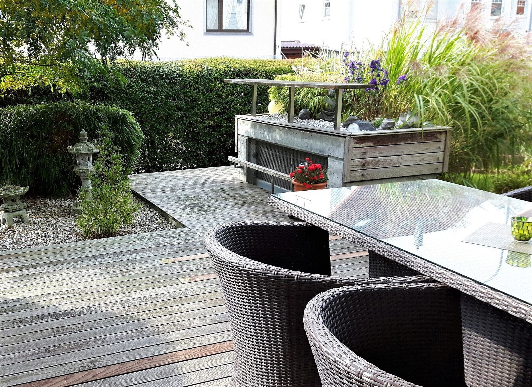 A wooden deck with a glass-top table and wicker chairs, surrounded by greenery and a stone lantern.