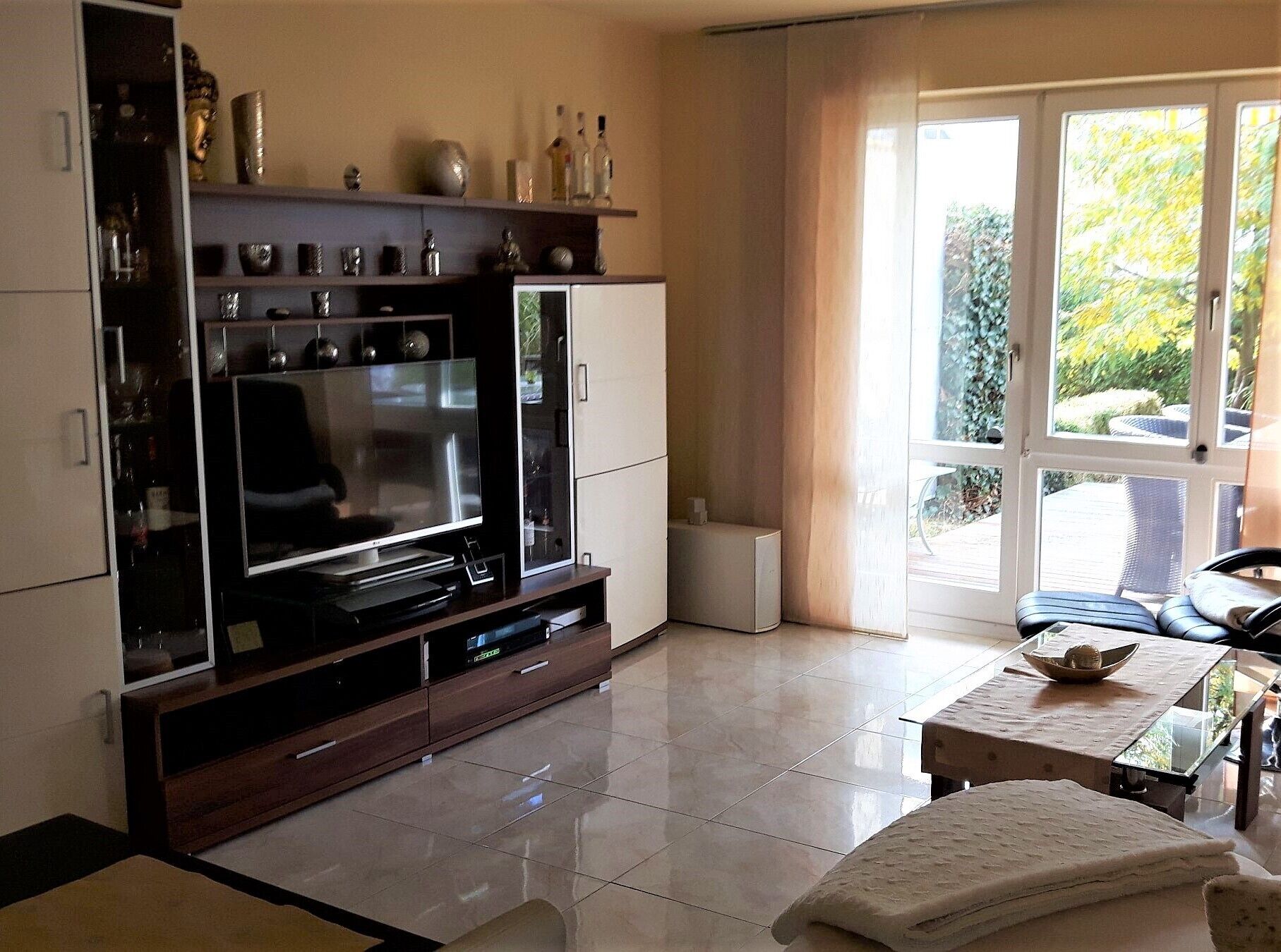 Ein Wohnzimmer mit einem großen Flachbildfernseher, einer Kommode mit Dekoration, einem Glastisch und Blick durch ein großes Fenster.