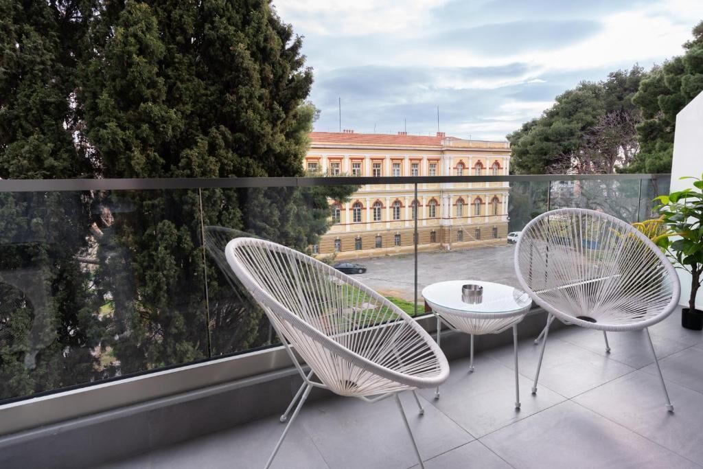 A balcony with two white wire chairs and a small table, overlooking a building with arched windows and a red roof.