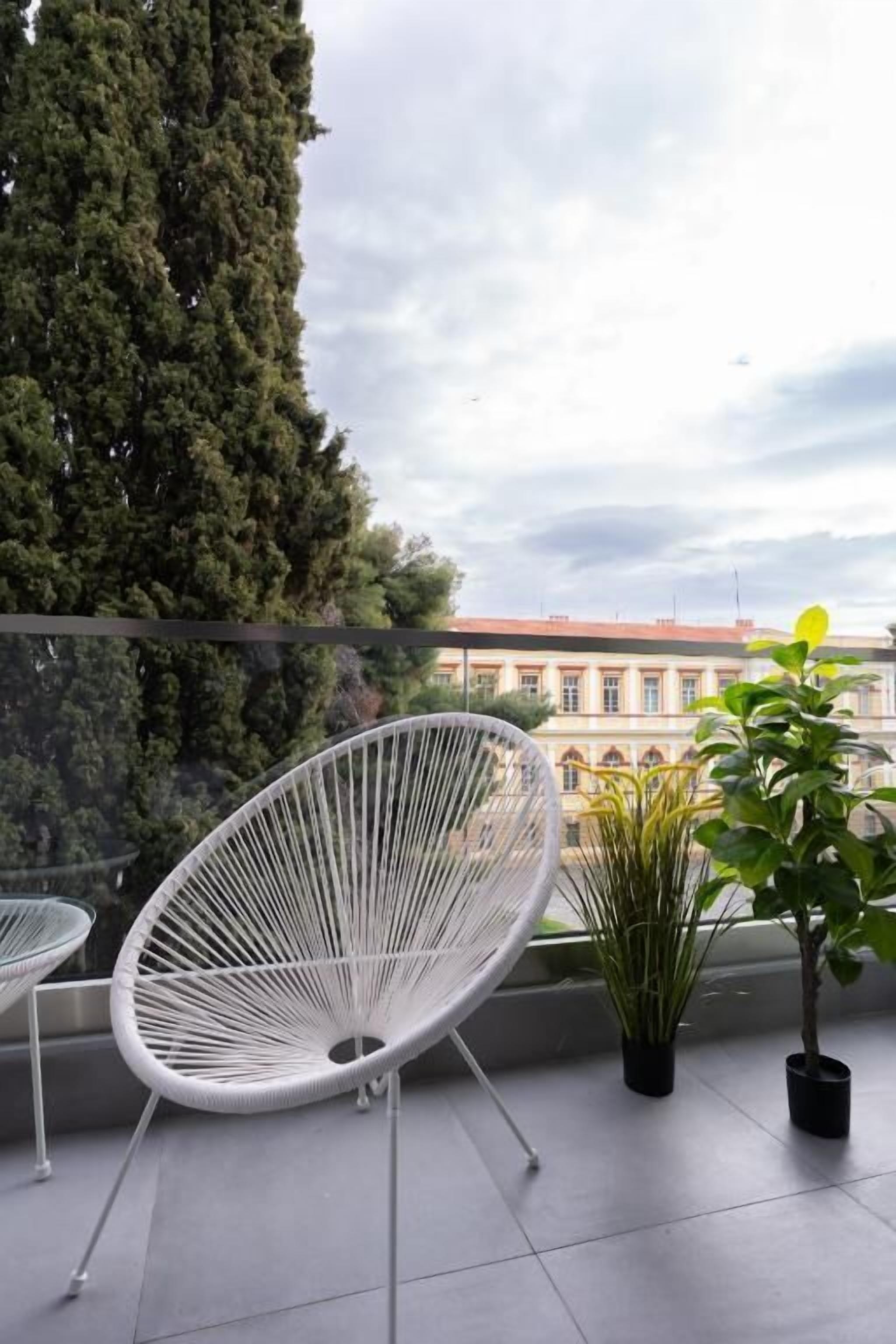 Een balkon met witte wicker stoelen, planten in potten en uitzicht op een gebouw met een rood dak.