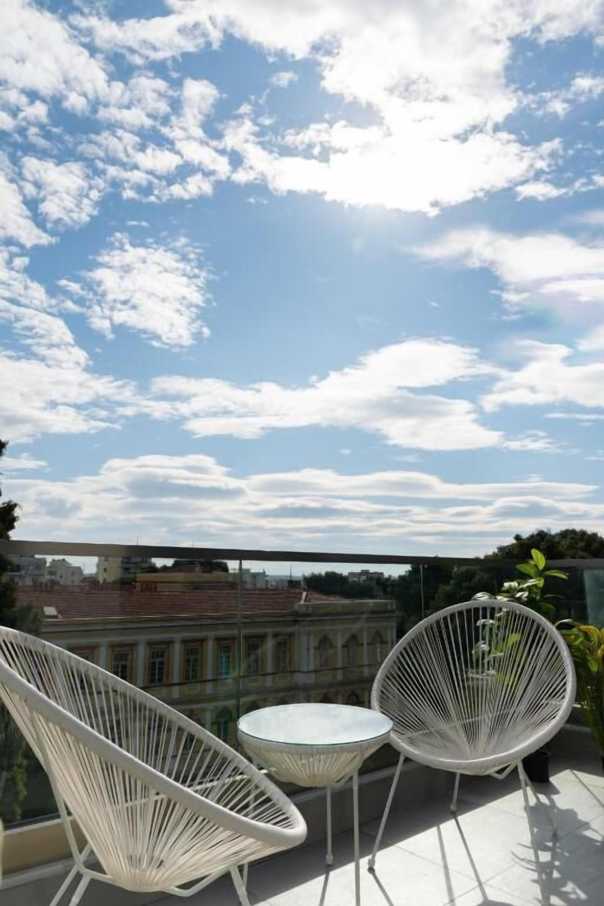 A balcony with two white wire-framed chairs and a small round table, overlooking a cityscape with historic buildings.