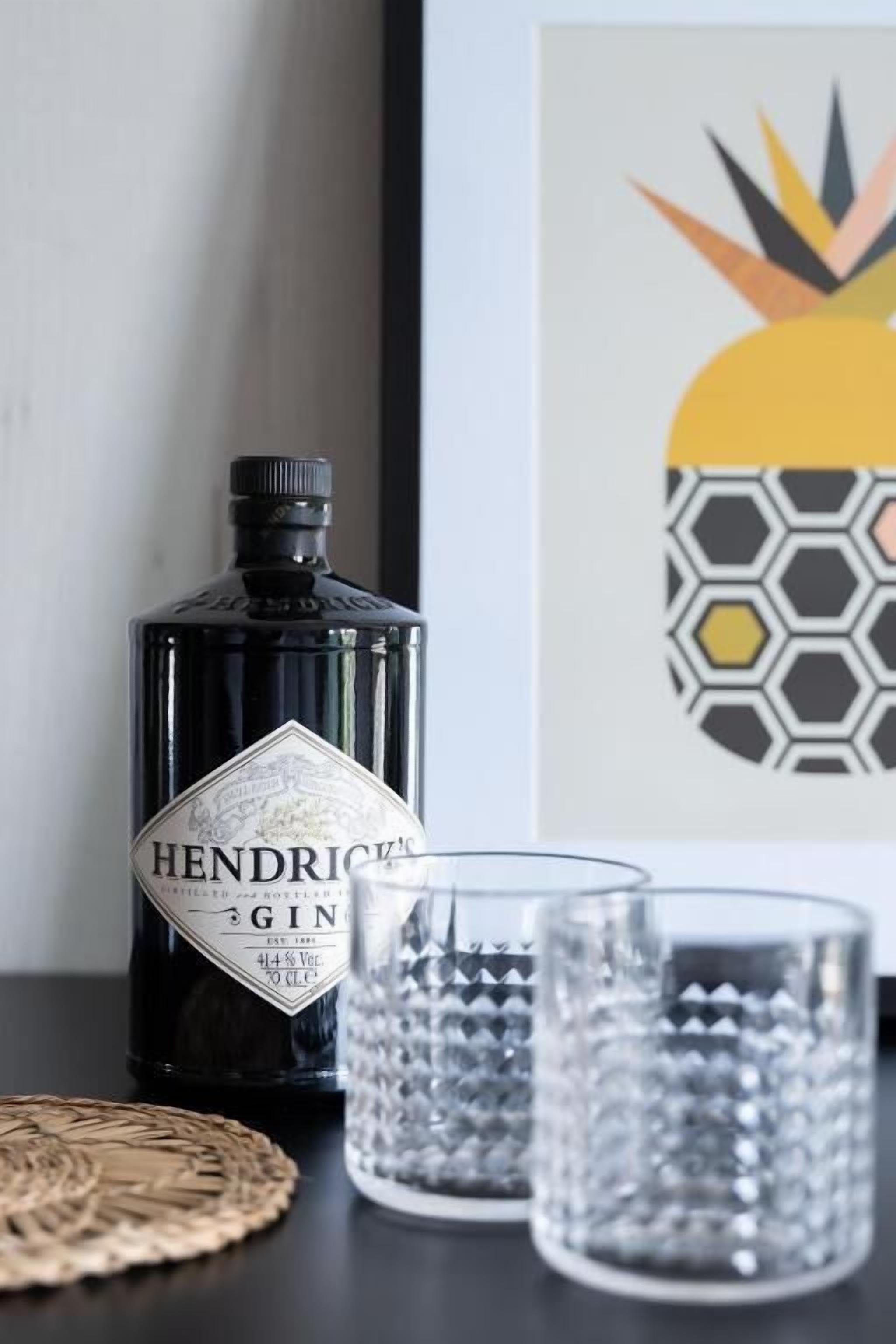 Una botella de ginebra Hendrick’s, dos vasos de vidrio y una obra de arte con piña en un marco sobre una mesa.