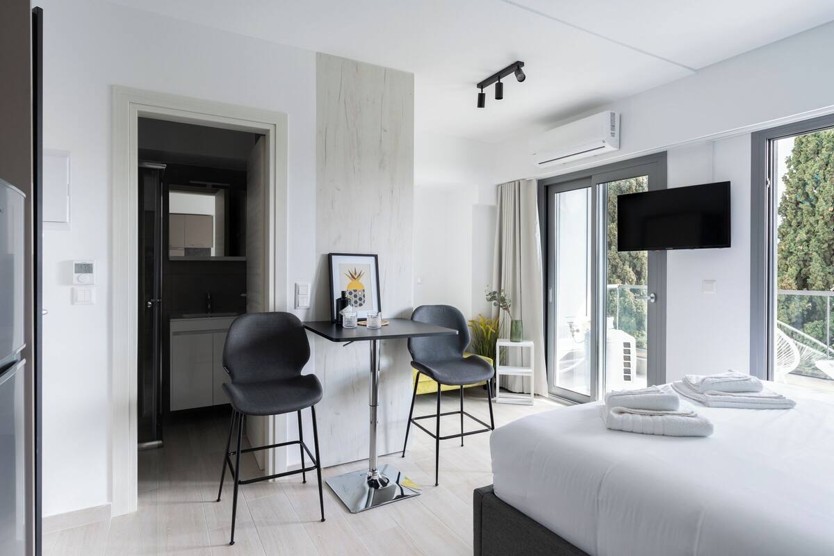 Een moderne hotelkamer met een groot bed, een bureau met twee stoelen, een televisie en een balkon met uitzicht op bomen.