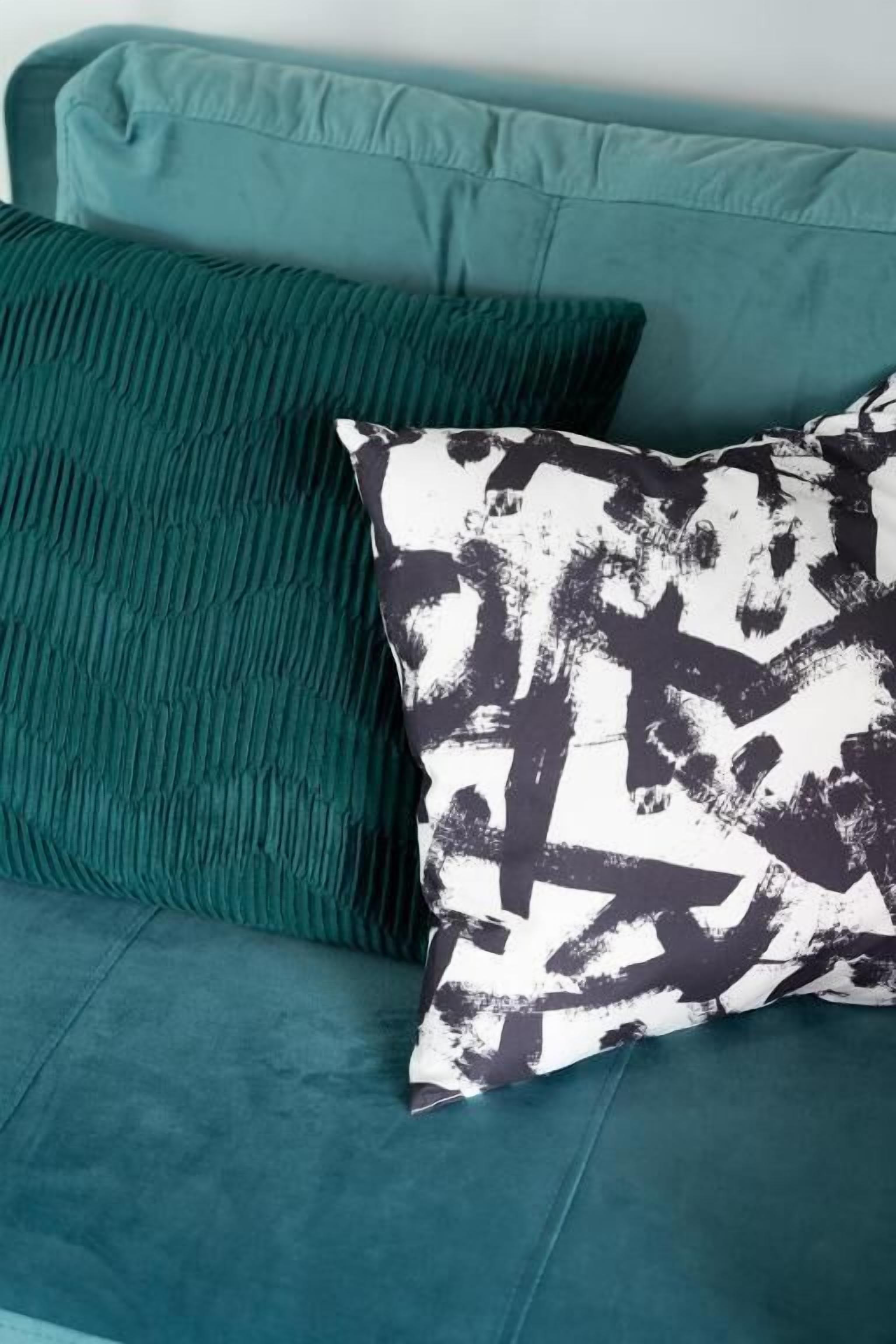 A teal sofa with two pillows, one with a textured pattern and the other with a black and white abstract design.