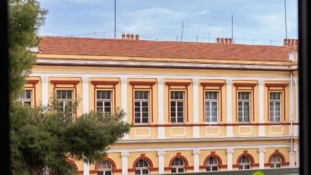 Vista de un edificio con tejado de tejas rojas y fachada amarilla, visible a través de una ventana. En primer plano, hay una silla blanca y una planta en maceta.