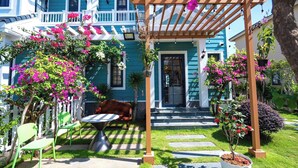 Una casa azul con un porche, una pérgola adornada con flores rosas y un jardín con sillas y una mesa.