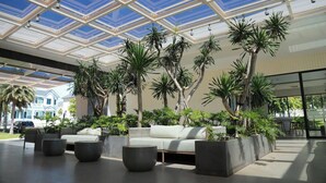 Un espacio interior amplio con techo de vidrio, un sofá blanco y numerosos maceteros con plantas tropicales.