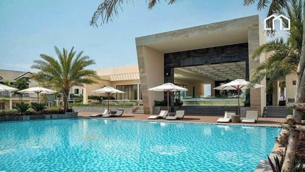Una piscina con sillas de descanso y sombrillas, rodeada de palmeras y un edificio moderno con una amplia entrada de vidrio.