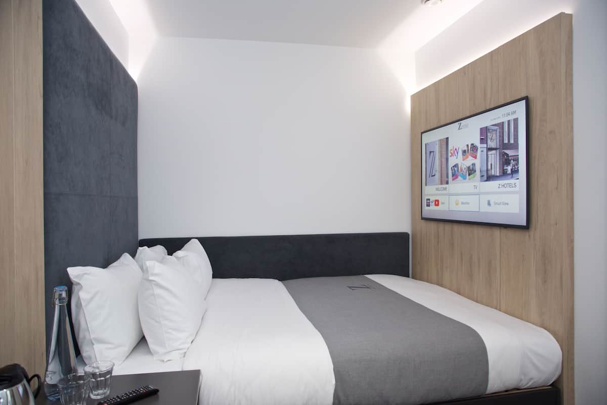 Una habitación de hotel moderna con una cama bien hecha, una mesita de noche con un control remoto y un cartel de bienvenida enmarcado en la pared.