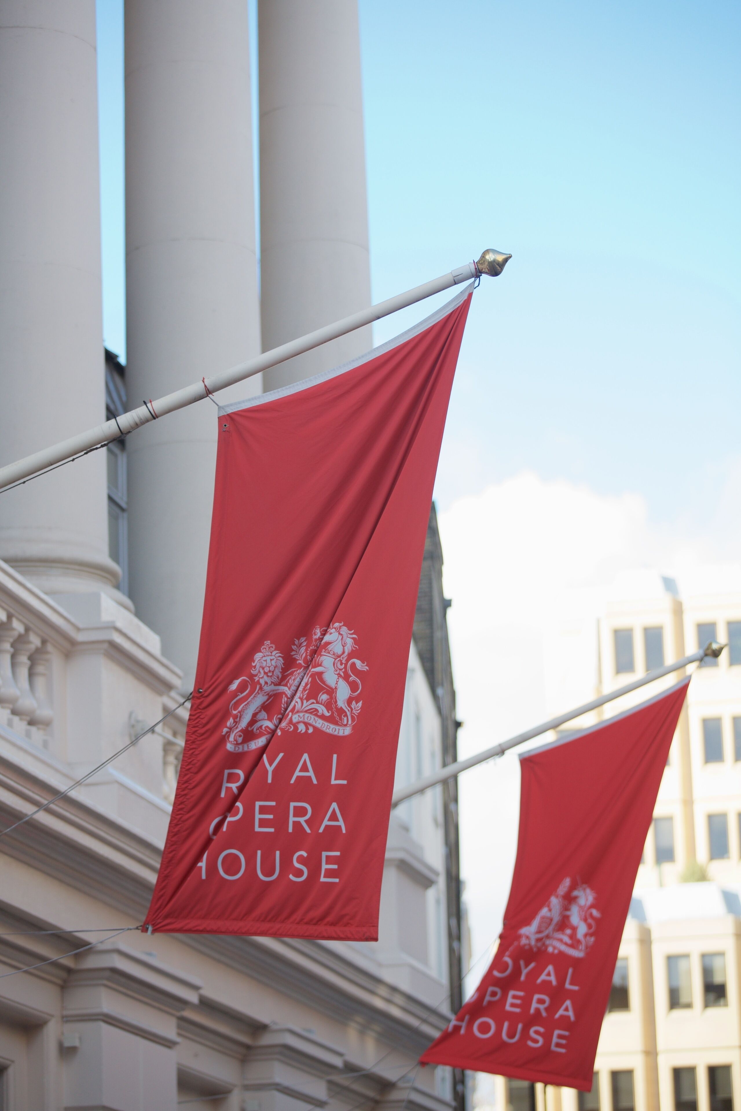 To røde flag med teksten "ROYAL OPERA HOUSE" og et løve-emblem, hængende på en flagstang foran en klassisk bygning med søjler.