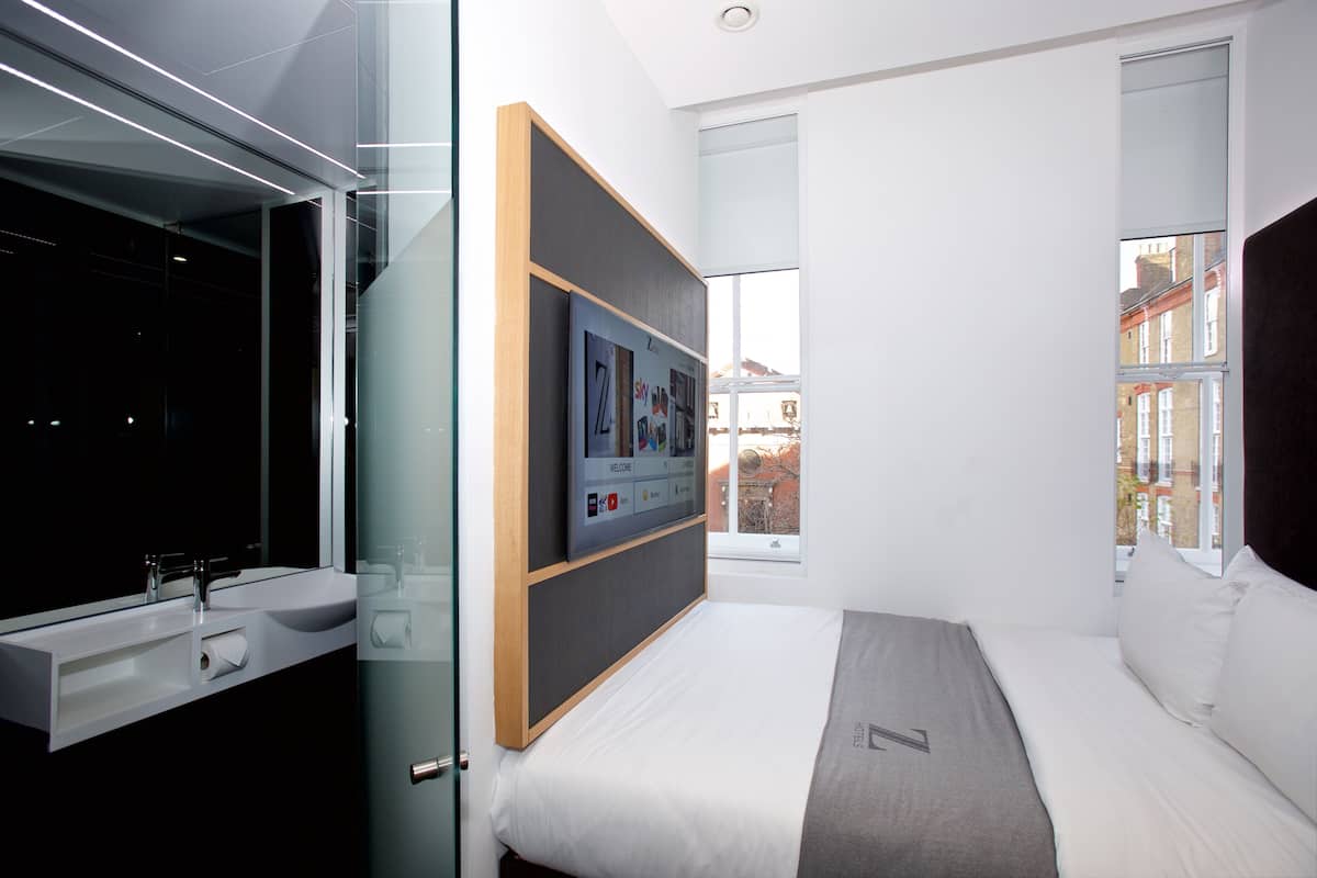 Habitación de hotel moderna con un ventanal grande, una cama, un televisor montado en la pared y un baño con lavamanos y espejo.