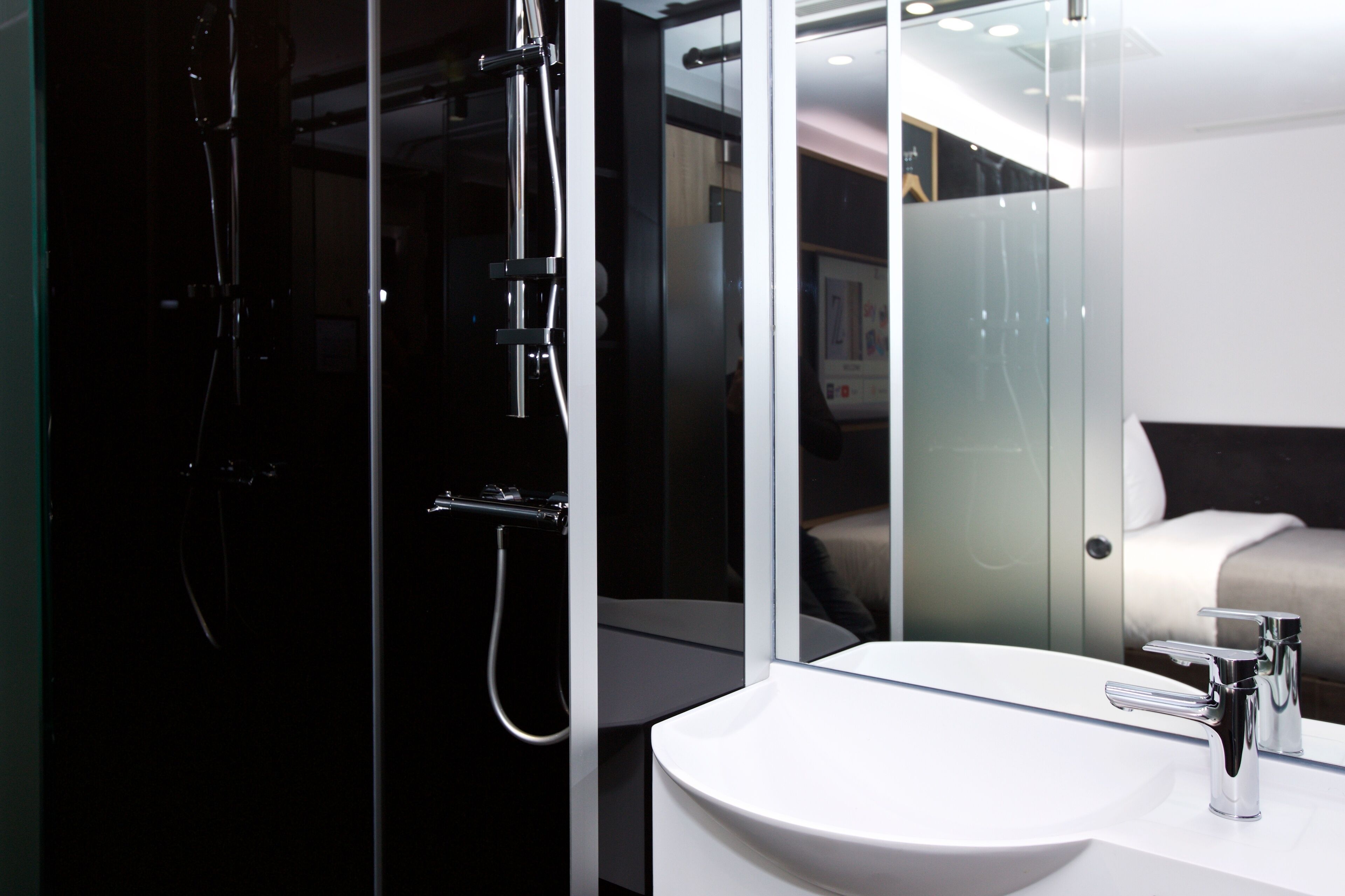 A modern bathroom with a glass-enclosed shower, a white sink, and a large mirror reflecting a bed and a wall-mounted TV.