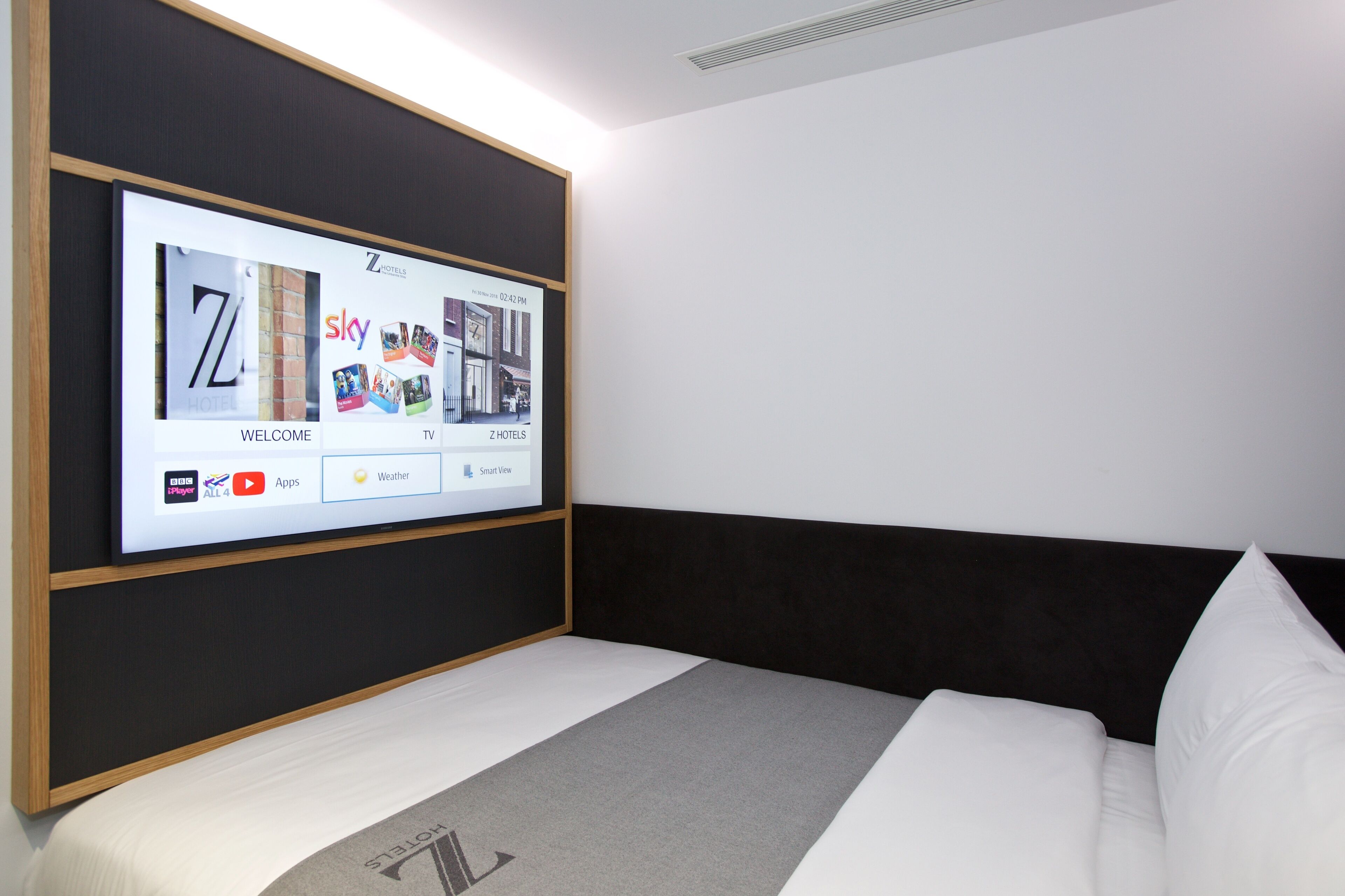 A modern hotel room with a large TV screen displaying a welcome screen, a neatly made bed, and a black headboard.