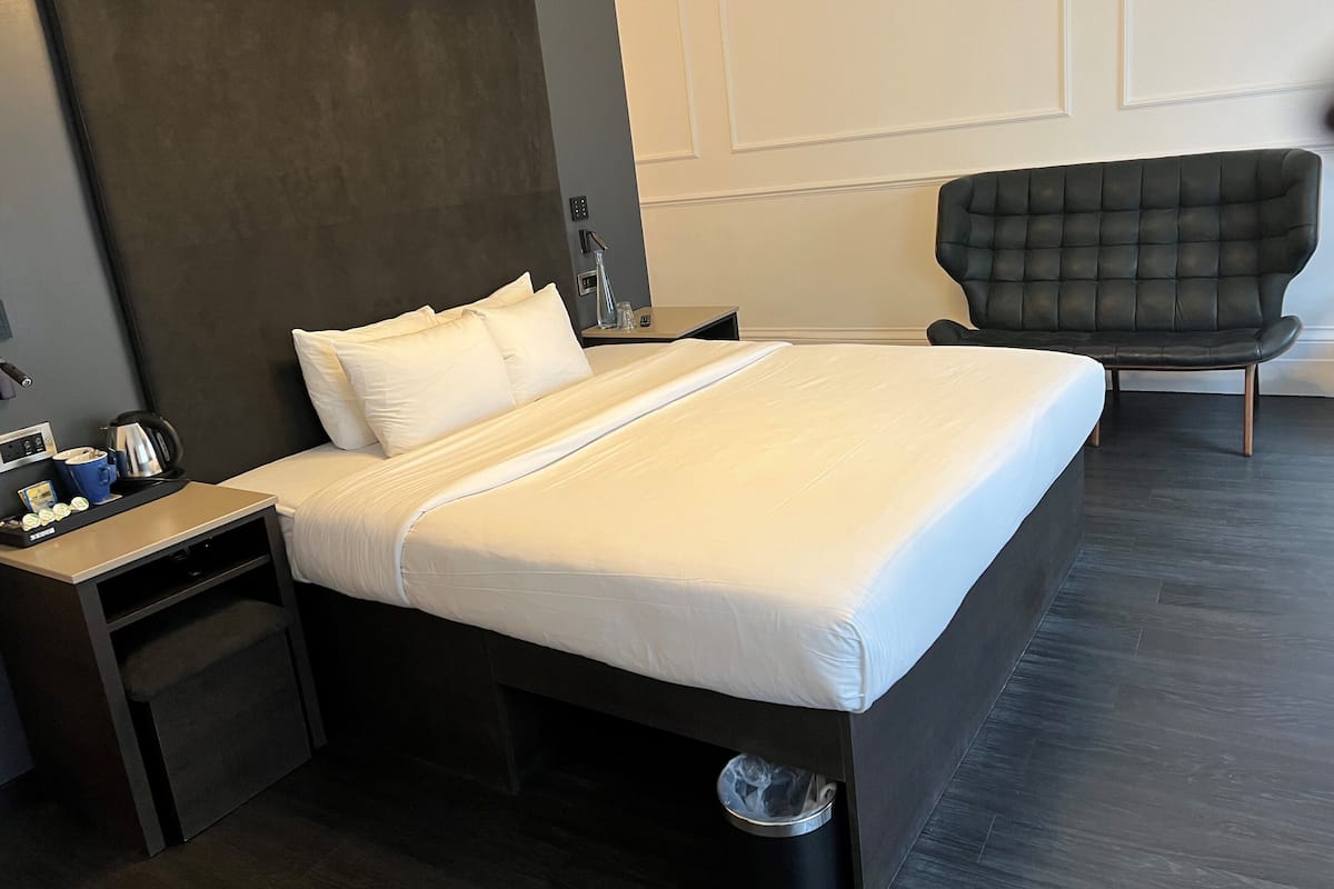 Una habitación de hotel moderna con una cama grande, un cabecero oscuro, una mesita de noche con hervidor, una silla y un cesto de basura.