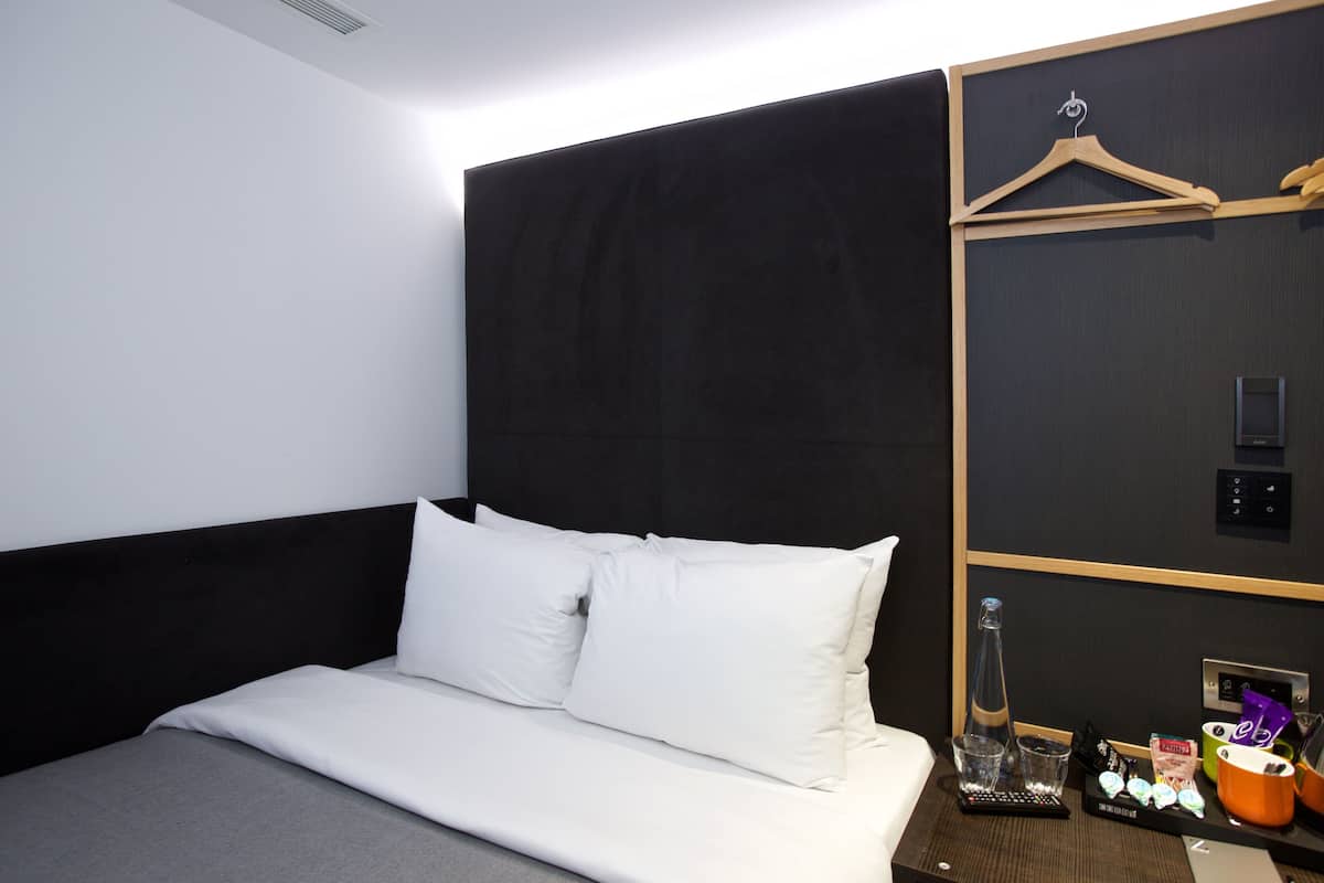 Una habitación de hotel moderna con una cama grande, un cabecero oscuro, una percha de madera, una botella, vasos y una bandeja con aperitivos.