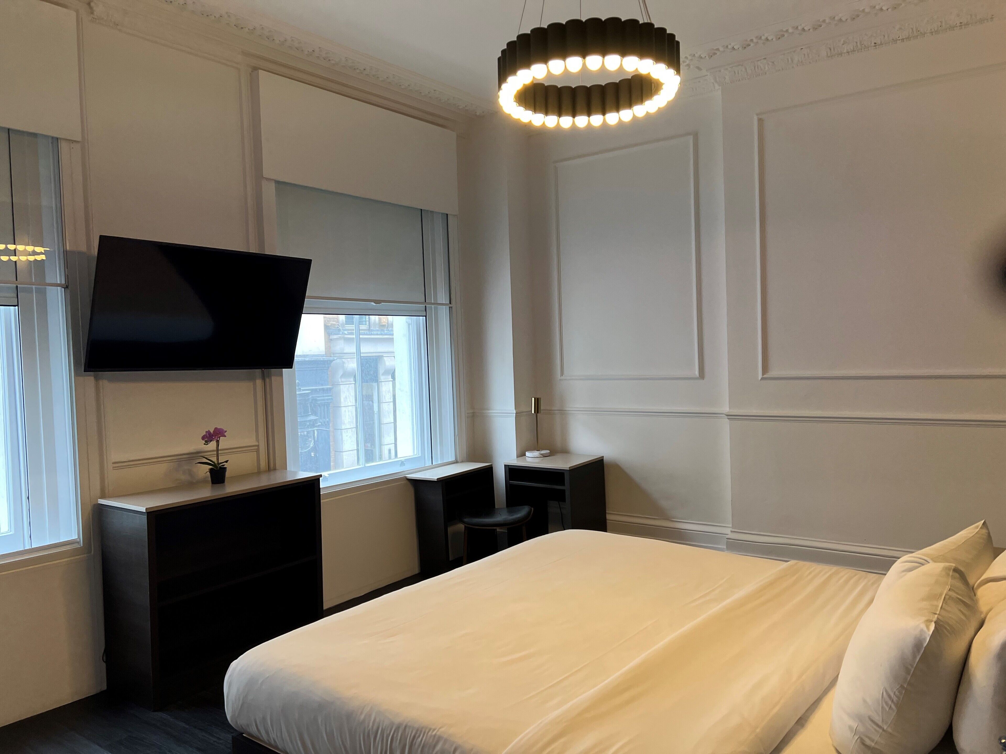 A hotel room with a bed, a desk, a TV, a window with a view of buildings, and a modern light fixture.