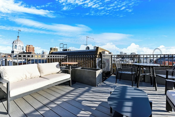A rooftop terrace with a sofa, dining table, and chairs, overlooking a cityscape with distinctive architecture and a Ferris wheel.