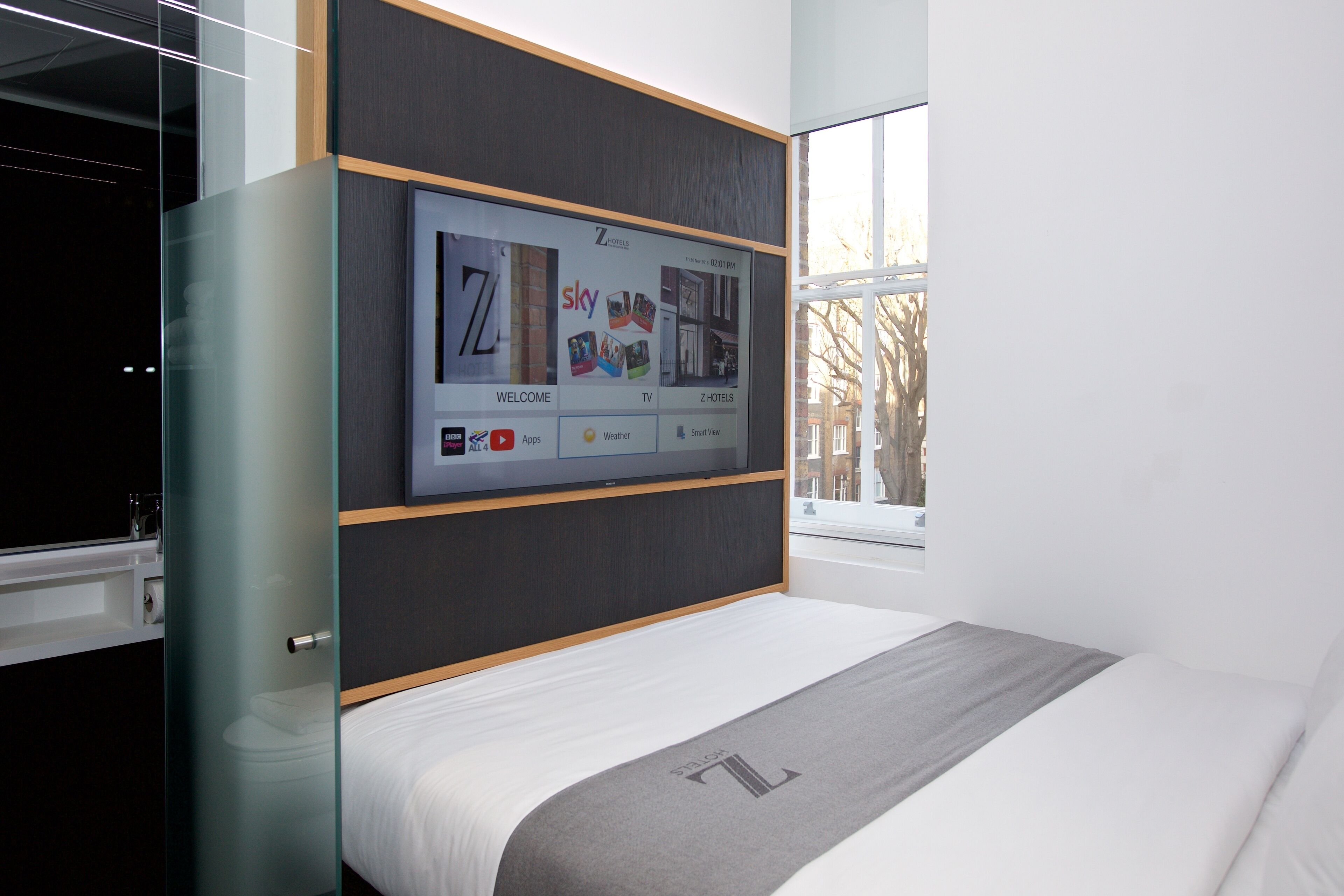 A modern hotel room with a large digital screen displaying a welcome screen, a bed with a grey headboard, and a window with a view of the outside.