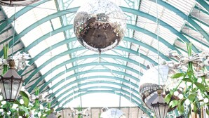 Una grande palla da discoteca sospesa sopra un soffitto in vetro in un ampio atrio ben illuminato, con travi strutturali verdi e lanterne pendenti.