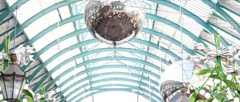A large disco ball hanging from a glass ceiling in a spacious, well-lit atrium with green structural beams and hanging lanterns.