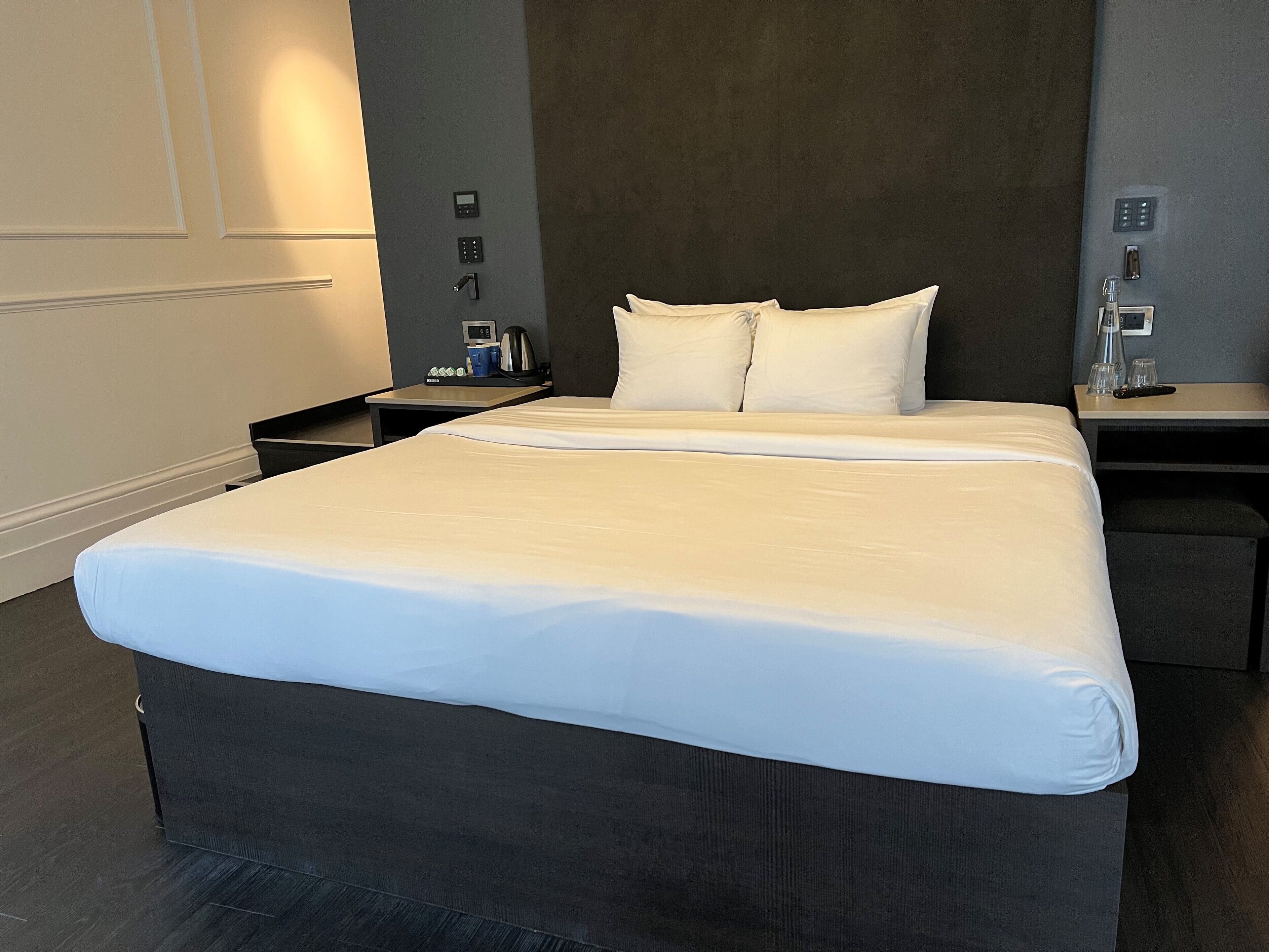 A hotel room with a large bed, a bedside table with a lamp, a power outlet, and a small table with a coffee maker.