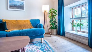 A living room with a blue sofa, a wooden floor lamp, a potted plant, and a view of the outdoors through a window with blue curtains.