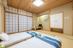 Une pièce de style japonais traditionnel, avec un sol recouvert de tatamis, deux lits avec une literie bleue et blanche, un bureau en bois, une télévision et des portes coulissantes.